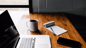 Bureau en bois avec un ordinateur portable ouvert, un carnet, un stylo, un smartphone et une tasse de café