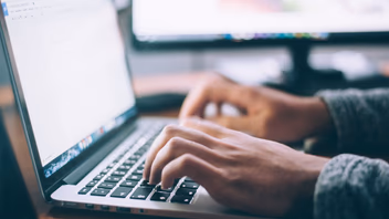 Mains tapant sur le clavier d’un ordinateur portable, un second écran allumé en arrière-plan