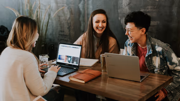 Trois personnes travaillent en riant autour d’une table avec leurs ordinateurs portables