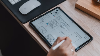 Main dessinant au stylet le wireframe d’une page web sur une tablette posée sur un bureau